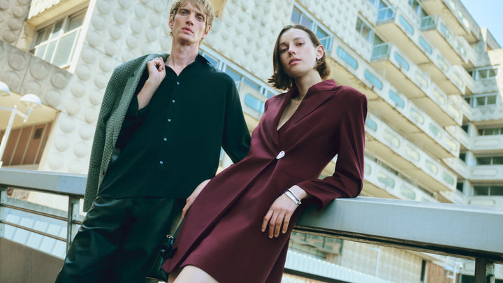 Two people in formal attire standing on a balcony with a city building in the background